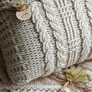 Puede incluir: Primer plano de una almohada de punto de color crema con un patr&oacute;n de punto de cable. La almohada tiene tres botones blancos y una etiqueta que dice "Made with Love".