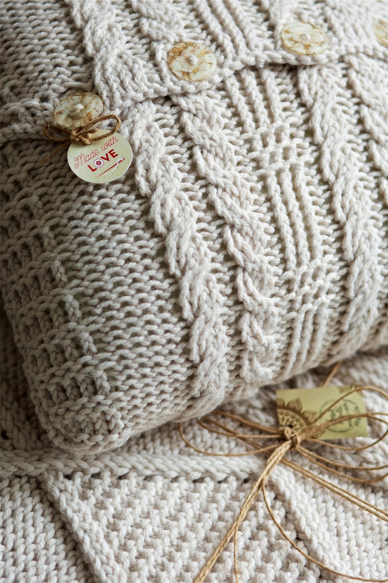 Puede incluir: Primer plano de una almohada de punto de color crema con un patr&oacute;n de punto de cable. La almohada tiene tres botones blancos y una etiqueta que dice "Made with Love".