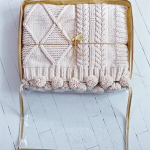 Puede incluir: Una manta de punto beige con un patr&oacute;n de rombos en un lado y un patr&oacute;n de punto trenzado en el otro. La manta est&aacute; doblada y atada con una cuerda y se guarda en una bolsa de pl&aacute;stico transparente con un asa marr&oacute;n.