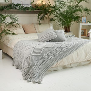 May include: A white bed with a gray knitted throw blanket and several decorative pillows. The bed is in a bedroom with a white floor and a potted plant.
