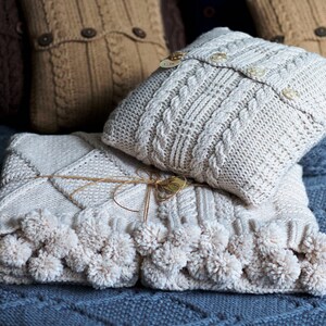 Puede incluir: Una manta de punto blanco con pompones en los bordes, doblada sobre una almohada de punto blanco con botones. La manta y la almohada est&aacute;n sobre una superficie de punto azul.