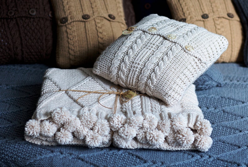Puede incluir: Una manta de punto blanco con pompones en los bordes, doblada sobre una almohada de punto blanco con botones. La manta y la almohada est&aacute;n sobre una superficie de punto azul.
