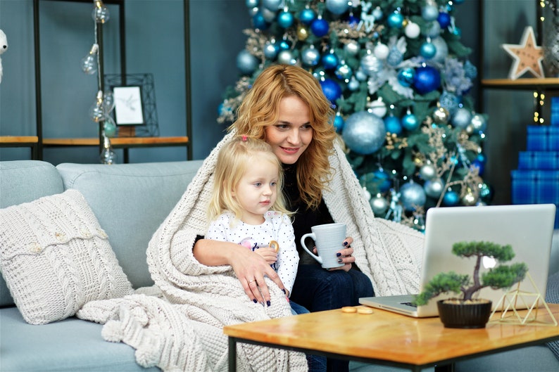 Puede incluir: Una mujer y una ni&ntilde;a peque&ntilde;a est&aacute;n sentadas en un sof&aacute; frente a un &aacute;rbol de Navidad, envueltas en una manta blanca, y mirando una computadora port&aacute;til. La mujer sostiene una taza y la ni&ntilde;a sostiene una galleta.