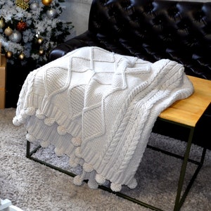 Puede incluir: Una manta de punto blanco con pompones en los bordes. La manta est&aacute; drapeada sobre una mesa de madera con un marco de metal negro. La mesa est&aacute; delante de un sof&aacute; de cuero negro.