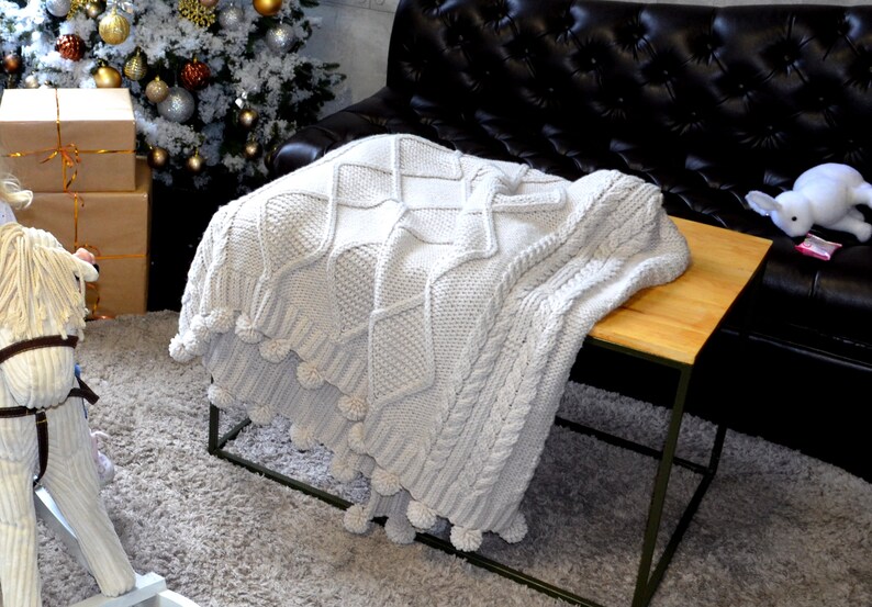 Puede incluir: Una manta de punto blanco con pompones en los bordes. La manta est&aacute; drapeada sobre una mesa de madera con un marco de metal negro. La mesa est&aacute; delante de un sof&aacute; de cuero negro.