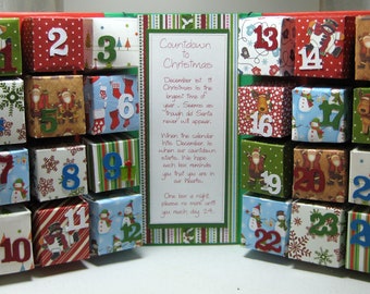 May include: A Christmas advent calendar with 24 numbered boxes, each decorated with a different festive pattern. The calendar is open to reveal a message about the countdown to Christmas.