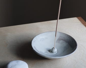 May include: A white ceramic incense burner with a wooden incense stick burning. Two smooth gray stones are in the foreground.