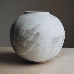 Puede incluir: Un jarrón de cerámica blanco con cuerpo redondo y un sutil patrón gris y blanco. El jarrón tiene una base ligeramente irregular.