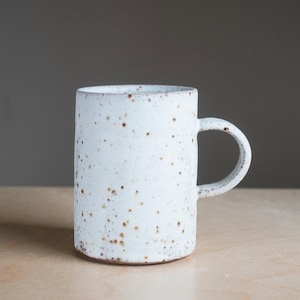 May include: A white ceramic mug with a speckled design and a curved handle. The mug has a cylindrical form and is decorated with small brown flecks. It rests on a light wooden surface.