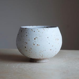 May include: A speckled white ceramic bowl with a rounded body and a small, circular base. The bowl has a wide opening and is set against a neutral background. The ceramic piece has brown speckles.