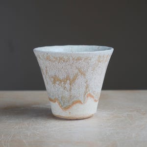 May include: A handmade ceramic cup with a textured, off-white exterior. The cup has a flared shape with a subtle orange and brown wave pattern near the base. The cup sits on a light-colored wooden surface.