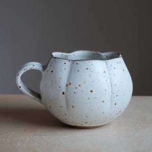Può includere: Una tazza in ceramica bianca con una forma arrotondata simile a una zucca e un bordo smerlato. La superficie presenta una consistenza maculata con piccoli punti marroni. Include un manico curvo sul lato sinistro e una base marrone.