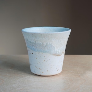 May include: A handmade ceramic cup with a flared rim. The cup features a speckled white base that transitions to a light blue and white band near the top. The interior is a light blue color.