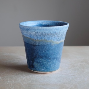 May include: A handmade ceramic tumbler with a gradient of blue hues. The top rim is a light blue, transitioning to a darker shade towards the base. The cup has a slightly flared shape and a textured surface.
