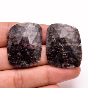 Könnte beinhalten: Zwei facettierte Edelsteine mit dunklem, gesprenkeltem Aussehen. Die Steine haben eine Mischung aus Dunkelgrau- und Schwarztönen mit kleinen Flecken von tiefem Rot. Die Form ist ein abgerundetes Rechteck mit polierter, reflektierender Oberfläche. Diese Steine könnten für Schmuck verwendet werden.