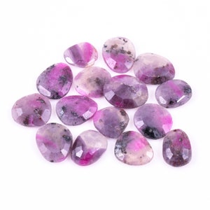 May include: A collection of faceted gemstones in shades of pink, purple, and grey. The stones have irregular shapes and varying sizes, with a polished, reflective surface. The gemstones are scattered on a white background.