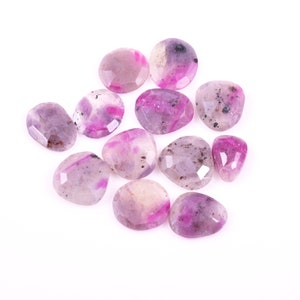 May include: A collection of faceted gemstones in shades of pink, purple, and white. The stones vary in shape, including round, oval, and teardrop. Some stones have dark speckles. The gemstones are scattered on a white background.
