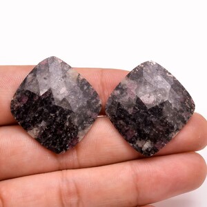 以下が含まれることがあります： 2つのファセット加工された正方形の宝石。石は、黒、グレー、ピンクのヒントを持つ、まだらの外観をしています。宝石は白い背景に対して手で持たれています。ジュエリー作りに適しています。