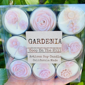 May include: A box of 9 Gardenia scented soy wax tea light candles. The candles are white with pink floral designs. The box has a label that reads "GARDENIA Coop On The Hill Artisan Soy Candle California Made."