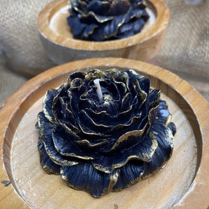 May include: Black rose-shaped candle with gold accents, set in a wooden tray. The candle has a white wick and detailed petal design. The second candle is visible in the background, also in a wooden tray. The candles are decorative.