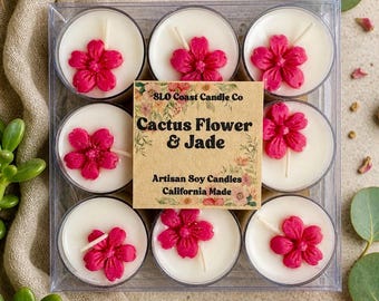 Op de afbeelding: Een doorzichtige doos met negen witte waxinelichtjes, elk versierd met een felroze bloem. Het label luidt "SLO Coast Candle Co Cactus Flower & Jade Artisan Soy Candles California Made."