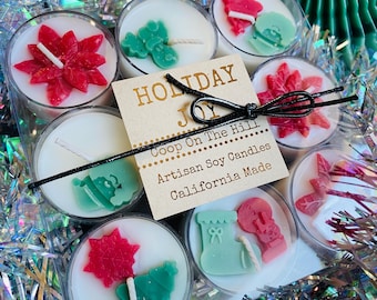 Op de afbeelding: Een set van acht feestelijke sojakaarsen in doorzichtige houders. Elke kaars heeft een witte wasbasis met decoratieve wasvormen in rood en groen, waaronder een poinsettia, kerstboom en laars. De set is vastgebonden met een zwart lint.