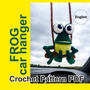May include: Crochet pattern PDF for a green frog car hanger. The frog is sitting on a brown swing and has big white eyes. The pattern is in English.