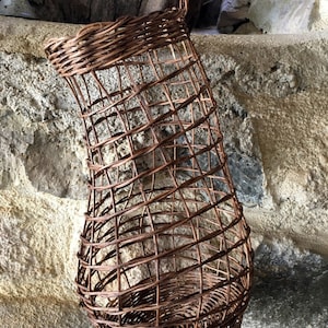 May include: A brown wicker basket with a unique, woven design. The basket is shaped like a vase and has a wide opening at the top. It is hanging from a hook on a stone wall.