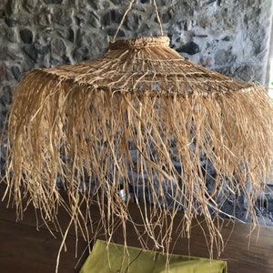 May include: A large, round, woven lampshade made of natural straw with a fringe of long, thin strands hanging down. The lampshade is hanging from the ceiling.