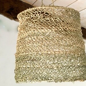 May include: A woven light fixture made of natural materials, with a light brown and green color scheme. The fixture is hanging from the ceiling and has a cylindrical shape.