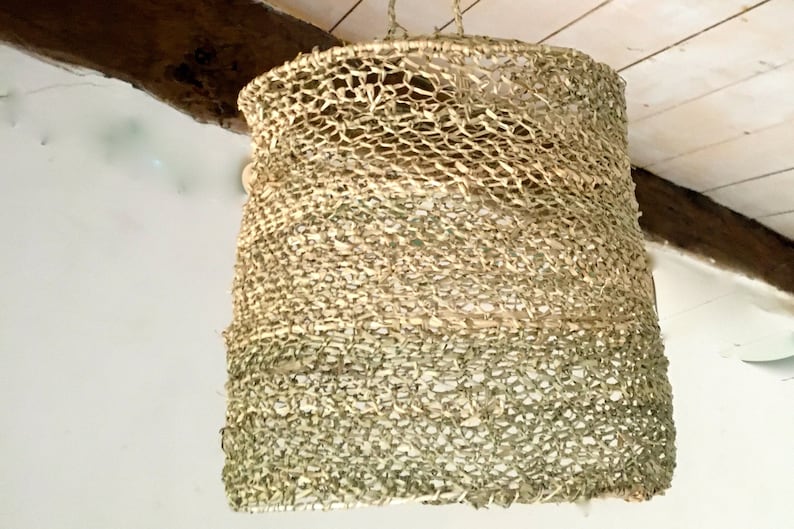 May include: A woven light fixture made of natural materials, with a light brown and green color scheme. The fixture is hanging from the ceiling and has a cylindrical shape.