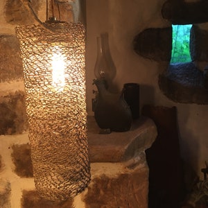 May include: A woven lampshade with a light bulb inside, hanging from the ceiling in a rustic setting. The lampshade is made of a natural material, possibly wicker or rattan, and has a warm, inviting glow.