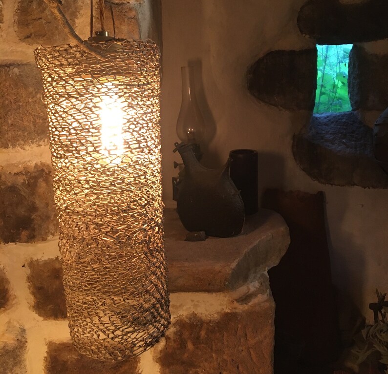 May include: A woven lampshade with a light bulb inside, hanging from the ceiling in a rustic setting. The lampshade is made of a natural material, possibly wicker or rattan, and has a warm, inviting glow.