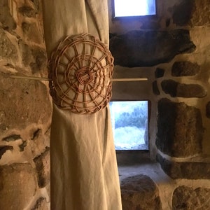 May include: A woven wicker circle with a wooden dowel attached to it. The circle is hanging from a beige curtain in front of a stone wall.