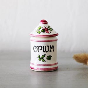 Puede incluir: Un tarro de cerámica blanco con tapa, con la palabra "Opium" en escritura negra. El tarro está decorado con detalles florales rosas y verdes y un borde rosa. El tarro mide aproximadamente 10 cm de alto.