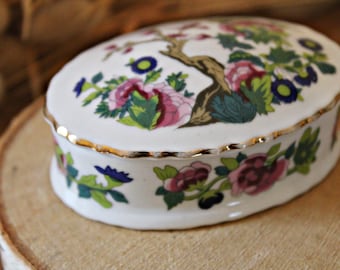 Może przedstawiać: Białe pudełko na drobiazgi z porcelany z pozłacanym wykończeniem i kwiatowym wzorem z różowymi, zielonymi i niebieskimi kwiatami. Pudełko ma w środku wzór gałęzi drzewa.