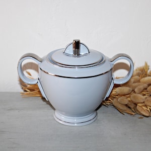 May include: A pale blue sugar bowl with a lid and two handles. The bowl has silver trim and a silver handle on the lid. The sugar bowl is on a grey surface.