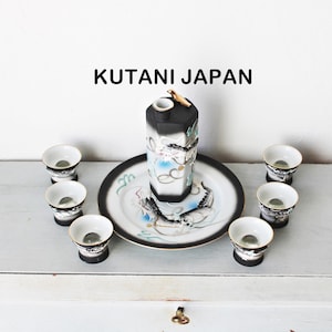 May include: A white ceramic mug with a black handle and the text "But First, Coffee" in a black, bold font. The mug is sitting on a wooden surface. The mug is approximately 4 inches tall.