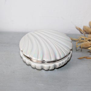 May include: A white, iridescent seashell-shaped trinket box with a silver-coloured rim. The box is open. The background features dried ornamental grass on a grey surface, creating a coastal theme.