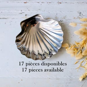 May include: A polished silver-toned metal dish shaped like a scallop shell with deep ridges and a reflective surface. The text 17 pièces disponibles 17 pieces available is printed below the shell, accompanied by a cluster of dried beige grasses.