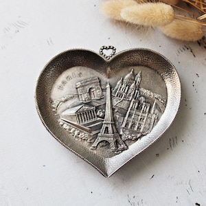 Könnte beinhalten: Herzförmige, silberfarbene Schmuckschale mit geprägten Pariser Wahrzeichen, darunter der Eiffelturm und der Arc de Triomphe. Das Wort "PARIS" steht oben. Dekorationsartikel.