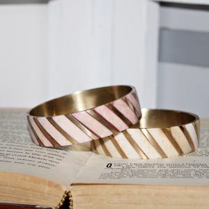 May include: Two brass bangles with a white and pink striped pattern. The bangles are stacked on top of each other.