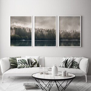 May include: A set of three framed prints depicting a misty forest scene with a lake in the foreground. The prints are in a modern style with a white frame and a muted colour palette.