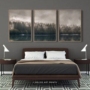 May include: Three framed prints depicting a misty forest scene with a lake in the foreground. The prints are hung on a grey wall above a brown leather bed with white bedding.