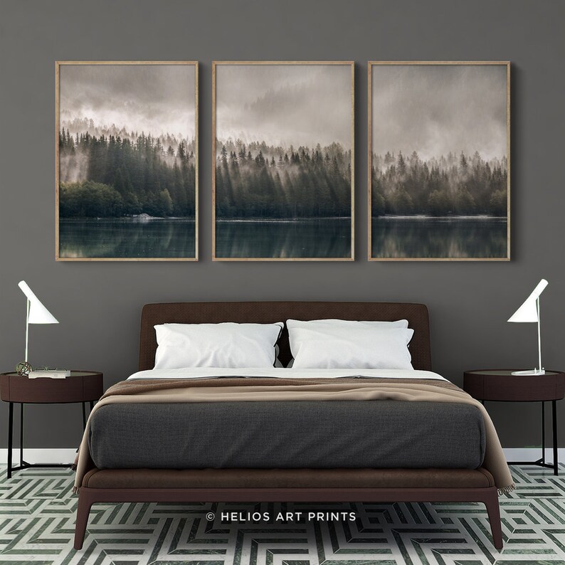 May include: Three framed prints depicting a misty forest scene with a lake in the foreground. The prints are hung on a grey wall above a brown leather bed with white bedding.