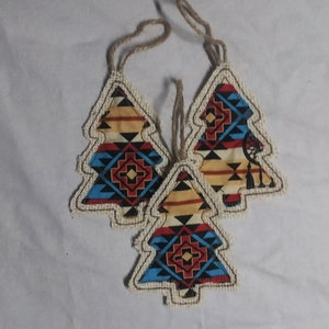 Op de afbeelding: Drie handgemaakte kerstboomversieringen met een geometrisch patroon geïnspireerd op de Amerikaanse inheemse cultuur in tinten rood, blauw, geel en zwart. De versieringen zijn gemaakt van stof en hebben een jute rand.