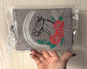 May include: A gray fabric pouch with a black horse head and red roses embroidered on it. The pouch has a horseshoe design around the horse head.