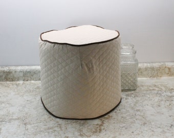 May include: A beige quilted cover with a brown trim for a kitchen appliance. The cover is sitting on a countertop.