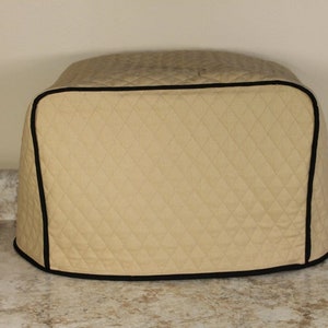 May include: Beige quilted toaster cover with a black trim. The cover is designed to fit a standard-sized toaster.