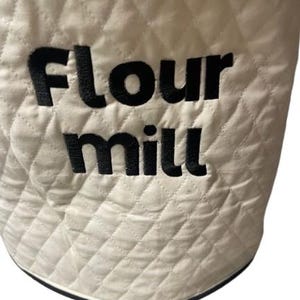 May include: Cream-colored quilted fabric with the words "Flour mill" embroidered in black. The fabric has a diamond pattern and a black trim along the bottom edge. The item appears to be a kitchen accessory.
